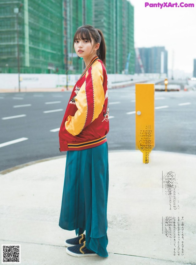 A woman in a red jacket and blue skirt standing on a sidewalk.